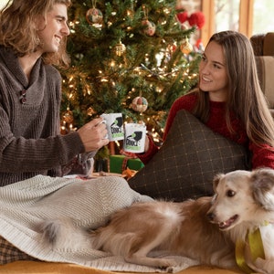 A young couple sit under a Christmas tree having a hot chocolate in matching white coffee mugs with Dink Responsibly funny Pickleball slogan on it.