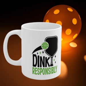 A dark background has a orange pickleball, that has lights glowing from it. In front of the light is a large white coffee mug with Dink Responsibly funny Pickleball slogan on it.