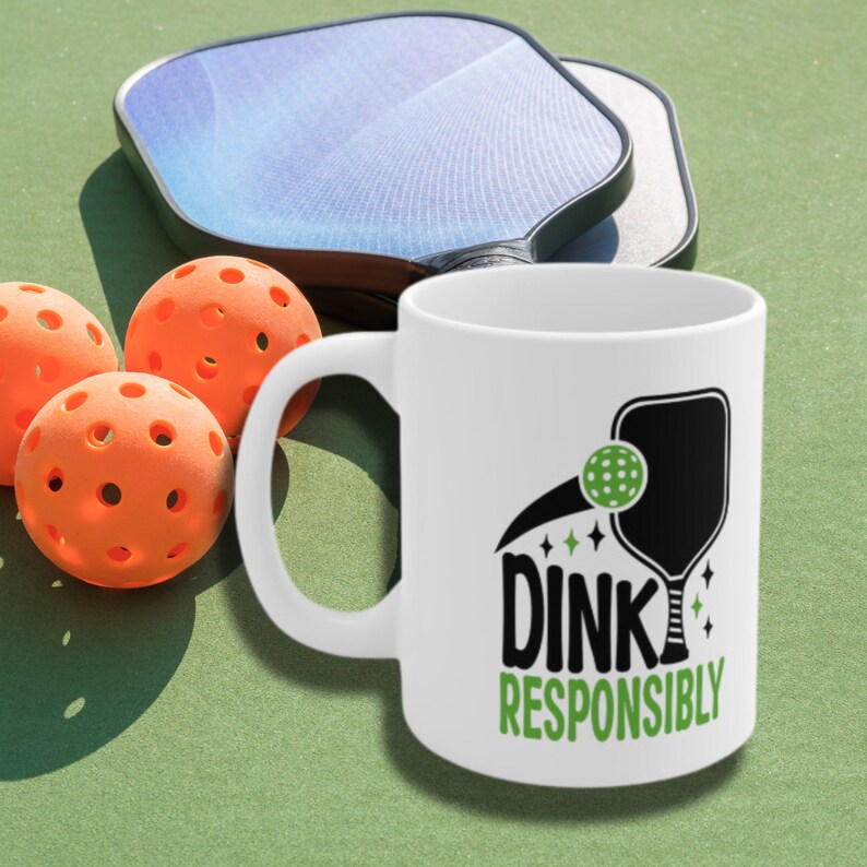 Two Pickleball paddles & three balls lie on a Pickleball court with a white coffee mug with Dink Responsibly funny Pickleball slogan on it, super imposed beside them.