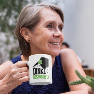 A middle aged woman holds a white coffee mug with Dink Responsibly funny Pickleball slogan on it.