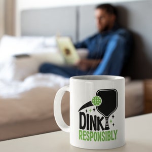Blurred back ground of a man in bed on a laptop and in the foreground is a white coffee mug with Dink Responsibly funny Pickleball slogan on it.