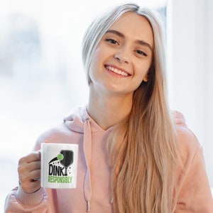 A young woman holds a white coffee mug with Dink Responsibly funny Pickleball slogan on it.