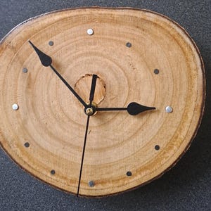 Könnte beinhalten: Eine Holzuhr mit schwarzen Zeigern und silbernen Akzenten. Das Zifferblatt der Uhr ist aus einer Baumscheibe mit sichtbaren Ringen.