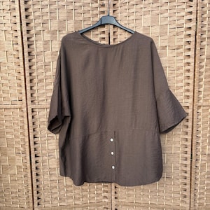 May include: A dark brown short-sleeved top with a wide, loose fit and a rounded neckline. The garment features a vertical placket on the lower front section adorned with four small white buttons, creating a subtle decorative detail on the fabric.