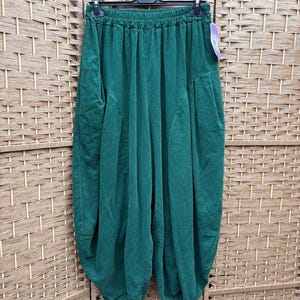 Pode incluir: Cal&ccedil;as verde-escuras com uma silhueta larga de estilo bal&atilde;o e cintura el&aacute;stica franzida. As cal&ccedil;as t&ecirc;m um corte descontra&iacute;do com detalhes de costura vertical na parte da frente e est&atilde;o penduradas num cabide de pl&aacute;stico preto.