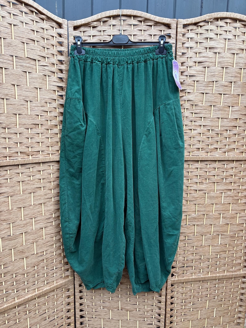 Pode incluir: Cal&ccedil;as verde-escuras com uma silhueta larga de estilo bal&atilde;o e cintura el&aacute;stica franzida. As cal&ccedil;as t&ecirc;m um corte descontra&iacute;do com detalhes de costura vertical na parte da frente e est&atilde;o penduradas num cabide de pl&aacute;stico preto.