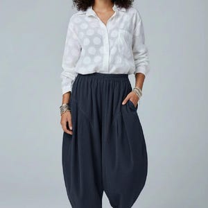 Pode incluir: Uma camisa branca de manga comprida com bot&otilde;es e um padr&atilde;o de grandes bolas brancas, combinada com cal&ccedil;as largas cinzento-carv&atilde;o escuro com cintura el&aacute;stica, bolsos laterais e uma silhueta volumosa, usadas com sapatilhas.