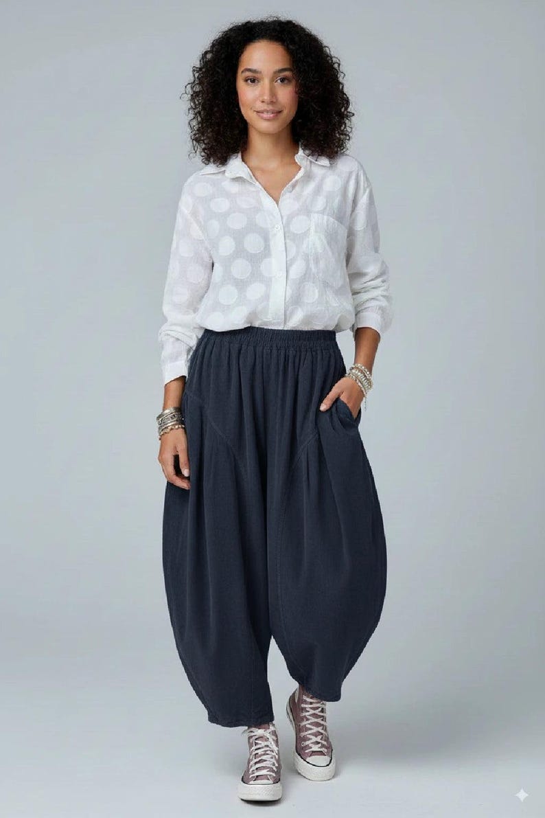 Pode incluir: Uma camisa branca de manga comprida com bot&otilde;es e um padr&atilde;o de grandes bolas brancas, combinada com cal&ccedil;as largas cinzento-carv&atilde;o escuro com cintura el&aacute;stica, bolsos laterais e uma silhueta volumosa, usadas com sapatilhas.