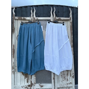 May include: Two pairs of wide-leg trousers hanging on black hangers. One pair is solid blue and the other is solid white. Both feature an elasticated waistband, a textured fabric finish, and a distinct diagonal seam detail across the front legs.
