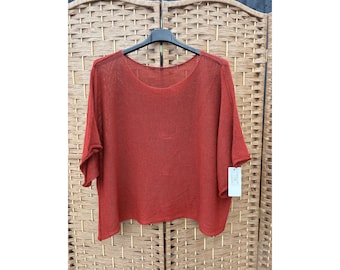 Pode incluir: Uma blusa de malha cor de ferrugem de corte largo, com um decote redondo amplo e mangas curtas e largas. A peça apresenta uma textura leve e semitransparente com uma bainha reta, pendurada num cabide preto com uma etiqueta branca anexada.