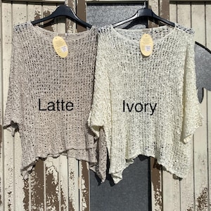 May include: Two crochet tops, one in ivory and one in latte, hanging on a wooden rack. The tops are made of a loose, open weave and have a relaxed fit.