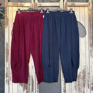 May include: Two pairs of casual trousers hang on a weathered wooden door. One pair is a deep burgundy, the other is a dark navy blue. Both have a gathered waist and a unique, voluminous design at the ankles.