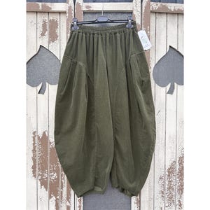 Pode incluir: Cal&ccedil;as largas verde-oliva com cintura el&aacute;stica alta, silhueta volumosa tipo bal&atilde;o e detalhes de costura vertical proeminentes na frente. A pe&ccedil;a &eacute; exibida num cabide de pl&aacute;stico preto com uma etiqueta branca.
