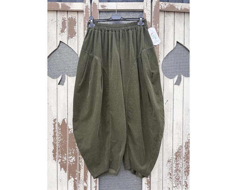 Pode incluir: Cal&ccedil;as largas verde-oliva com cintura el&aacute;stica alta, silhueta volumosa tipo bal&atilde;o e detalhes de costura vertical proeminentes na frente. A pe&ccedil;a &eacute; exibida num cabide de pl&aacute;stico preto com uma etiqueta branca.