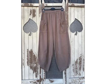 Pode incluir: Calças de cor taupe sólida com uma silhueta relaxada de perna larga, cintura elástica e dois bolsos grandes e proeminentes na parte frontal. As calças têm uma bainha afunilada e estão expostas num cabide de plástico preto.