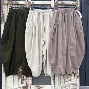 Pode incluir: Tr&ecirc;s cal&ccedil;as largas penduradas em cabides de pl&aacute;stico preto. As cal&ccedil;as est&atilde;o dispon&iacute;veis em verde-azeitona, branco-sujo e castanho-claro. Cada par apresenta uma cintura el&aacute;stica, bolsos laterais e uma silhueta volumosa e arredondada.