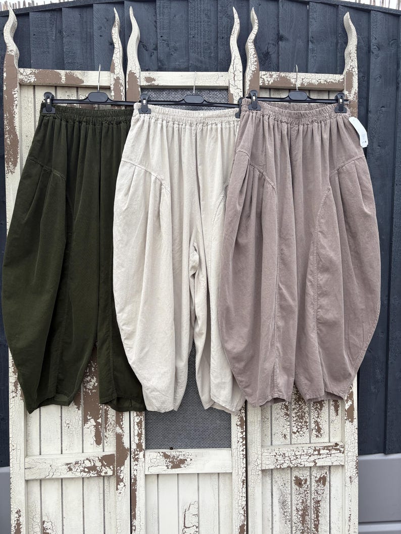 Pode incluir: Tr&ecirc;s cal&ccedil;as largas penduradas em cabides de pl&aacute;stico preto. As cal&ccedil;as est&atilde;o dispon&iacute;veis em verde-azeitona, branco-sujo e castanho-claro. Cada par apresenta uma cintura el&aacute;stica, bolsos laterais e uma silhueta volumosa e arredondada.