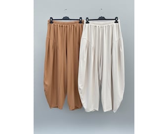 Pode incluir: Dois pares de calças de corte largo pendurados em cabides de plástico preto. Um par é de cor castanho-claro sólido e o outro é de cor branco-sujo. Ambas as calças apresentam cintura elástica, corte de cintura subida e pernas largas.