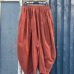 Pode incluir: Cal&ccedil;as de veludo cotel&ecirc; na cor ferrugem com cintura el&aacute;stica alta, silhueta volumosa de perna bal&atilde;o e bolsos laterais profundos. As cal&ccedil;as est&atilde;o penduradas num cabide preto, destacando o tecido texturizado e o corte largo da pe&ccedil;a.