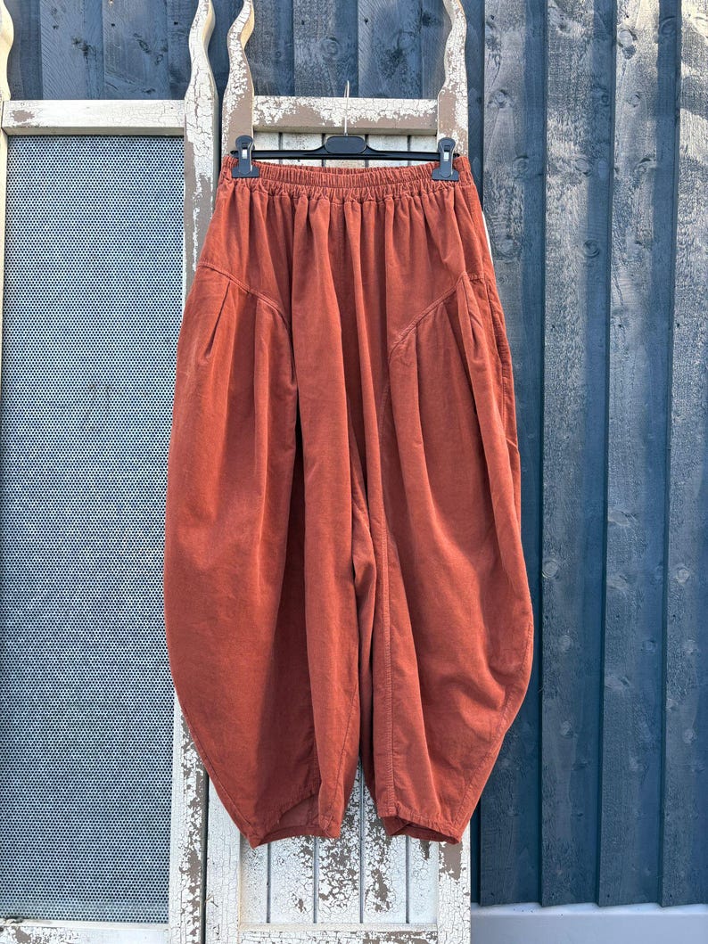 Pode incluir: Cal&ccedil;as de veludo cotel&ecirc; na cor ferrugem com cintura el&aacute;stica alta, silhueta volumosa de perna bal&atilde;o e bolsos laterais profundos. As cal&ccedil;as est&atilde;o penduradas num cabide preto, destacando o tecido texturizado e o corte largo da pe&ccedil;a.