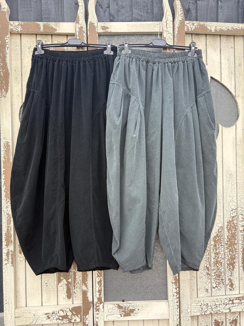Pode incluir: Dois pares de cal&ccedil;as de corte largo pendurados em cabides de pl&aacute;stico preto. Um par &eacute; preto s&oacute;lido e o outro &eacute; de um cinzento escuro suave. Ambos apresentam cintura el&aacute;stica, bolsos laterais profundos e uma silhueta de perna arredondada.