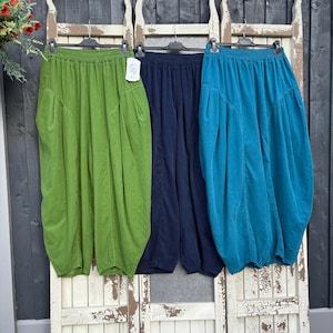Pode incluir: Tr&ecirc;s cal&ccedil;as largas com cinturas el&aacute;sticas, cada uma numa cor distinta: verde-lima, azul-marinho escuro e azul-petr&oacute;leo. Estas cal&ccedil;as de perna larga t&ecirc;m uma silhueta franzida com bolsos laterais e est&atilde;o penduradas em cabides pretos.