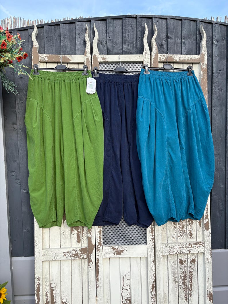 Pode incluir: Tr&ecirc;s cal&ccedil;as largas com cinturas el&aacute;sticas, cada uma numa cor distinta: verde-lima, azul-marinho escuro e azul-petr&oacute;leo. Estas cal&ccedil;as de perna larga t&ecirc;m uma silhueta franzida com bolsos laterais e est&atilde;o penduradas em cabides pretos.