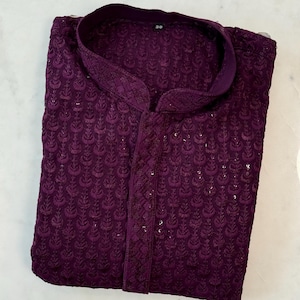 Pu&ograve; includere: Una camicia kurta viola scuro piegata con ricami floreali intricati. La camicia presenta un colletto alla coreana e una paramontura abbottonata. Il tessuto ha una lucentezza sottile e il design &egrave; formale ed elegante.
