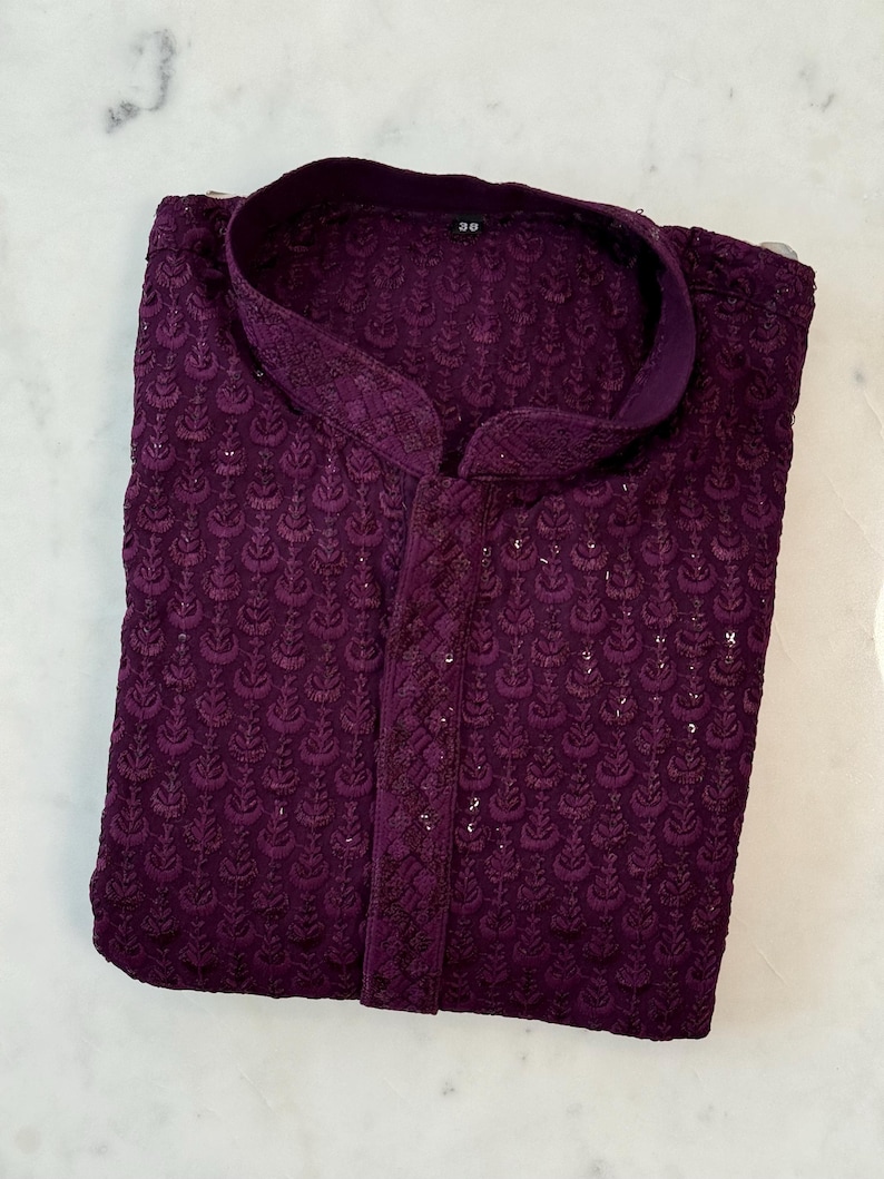 Pu&ograve; includere: Una camicia kurta viola scuro piegata con ricami floreali intricati. La camicia presenta un colletto alla coreana e una paramontura abbottonata. Il tessuto ha una lucentezza sottile e il design &egrave; formale ed elegante.
