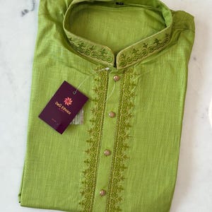 May include: A lime green kurta shirt with detailed embroidery on the collar and front. The shirt features a mandarin collar and gold buttons. A tag from "Desi Ethnics" is attached to the garment.