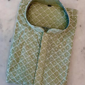 Pu&ograve; includere: Una camicia verde piegata con un motivo geometrico bianco. La camicia ha un colletto alla coreana e una chiusura con bottoni. Il tessuto sembra leggero, adatto al clima caldo. La camicia &egrave; ben piegata su una superficie di marmo.