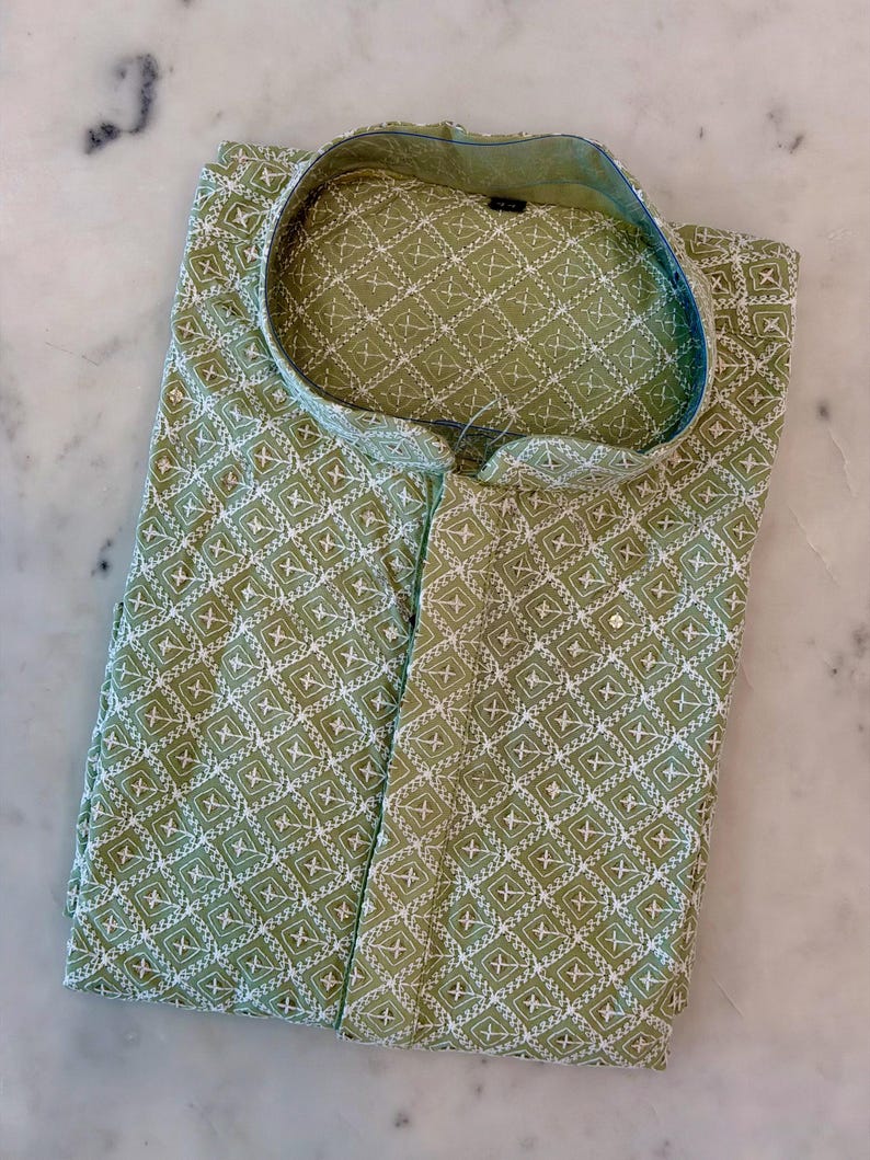 Pu&ograve; includere: Una camicia verde piegata con un motivo geometrico bianco. La camicia ha un colletto alla coreana e una chiusura con bottoni. Il tessuto sembra leggero, adatto al clima caldo. La camicia &egrave; ben piegata su una superficie di marmo.