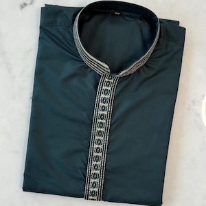 May include: A dark teal tunic with a mandarin collar. The neckline and placket are decorated with a trim featuring a repeating geometric pattern in green and gold. The shirt is neatly folded on a white marble surface.
