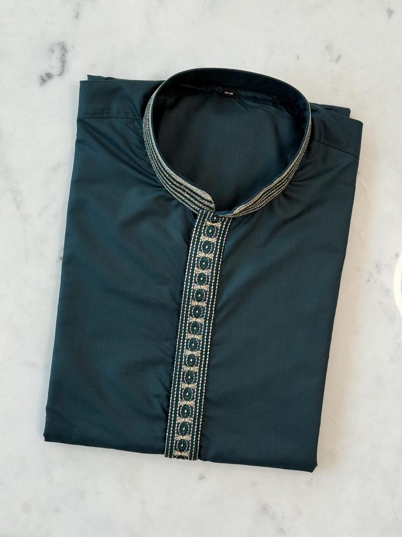 May include: A dark teal tunic with a mandarin collar. The neckline and placket are decorated with a trim featuring a repeating geometric pattern in green and gold. The shirt is neatly folded on a white marble surface.