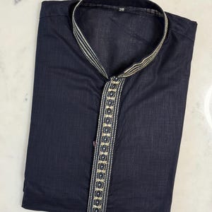 May include: Navy blue kurta shirt with a mandarin collar and decorative trim. The trim features a gold and silver pattern with circular accents. The shirt is folded neatly, showing the front and collar details. The number "38" is visible on the collar.