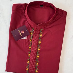 May include: A maroon kurta shirt with a mandarin collar and detailed embroidery along the placket. The embroidery includes green, yellow, and white floral designs. A tag from "DeQ Ethnics" is attached.