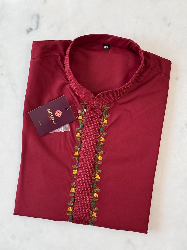 May include: A maroon kurta shirt with a mandarin collar and detailed embroidery along the placket. The embroidery includes green, yellow, and white floral designs. A tag from "DeQ Ethnics" is attached.