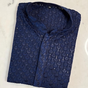 Pu&ograve; includere: Camicia kurta blu navy con dettagli ricamati. La camicia presenta un colletto alla coreana e una chiusura frontale con bottoni. Il tessuto ha una leggera lucentezza e il ricamo crea una superficie strutturata. L'indumento &egrave; piegato con cura.