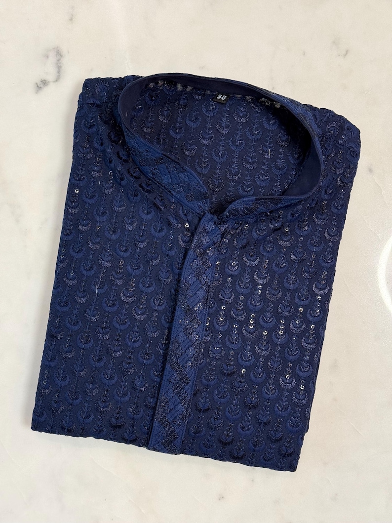Pu&ograve; includere: Camicia kurta blu navy con dettagli ricamati. La camicia presenta un colletto alla coreana e una chiusura frontale con bottoni. Il tessuto ha una leggera lucentezza e il ricamo crea una superficie strutturata. L'indumento &egrave; piegato con cura.