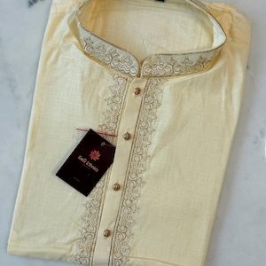 May include: Cream-coloured kurta shirt with gold embroidery on the collar and front. The shirt features a mandarin collar and gold buttons. A tag from "DeS Ethnics" is attached.