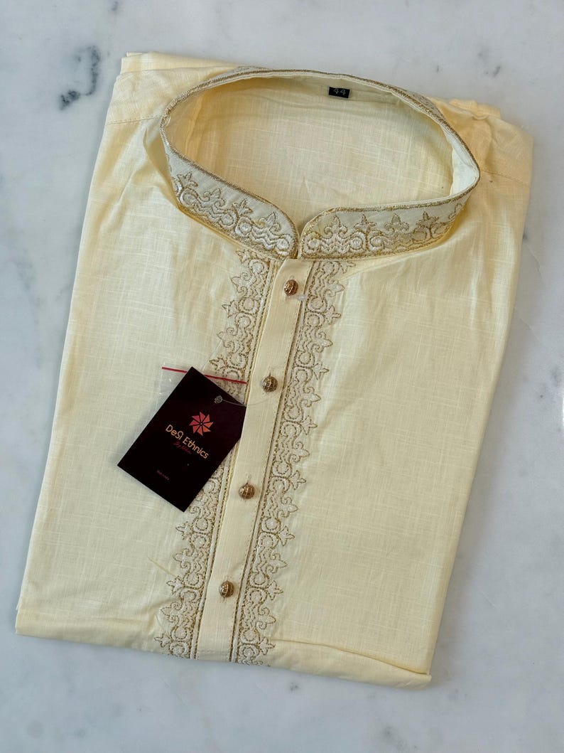 May include: Cream-coloured kurta shirt with gold embroidery on the collar and front. The shirt features a mandarin collar and gold buttons. A tag from "DeS Ethnics" is attached.