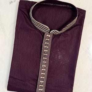 May include: A folded, dark burgundy kurta shirt with a mandarin collar and a decorative embroidered trim. The trim features a repeating pattern of gold and burgundy accents. The shirt is made of a lightweight fabric.