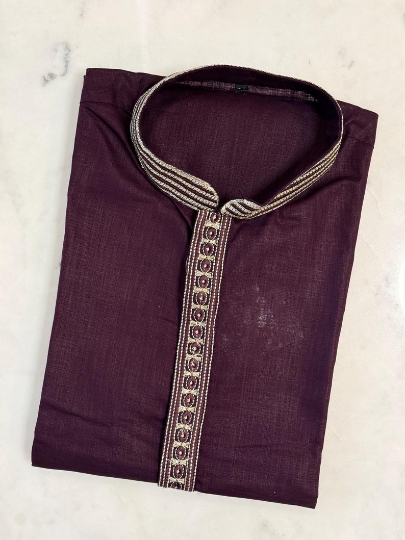 May include: A folded, dark burgundy kurta shirt with a mandarin collar and a decorative embroidered trim. The trim features a repeating pattern of gold and burgundy accents. The shirt is made of a lightweight fabric.