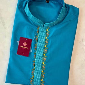 May include: A turquoise kurta shirt with a mandarin collar and embroidered detailing. The shirt features a vertical embroidered panel with floral accents. A tag from "DeSi Ethnics" is attached. The size tag reads "42".