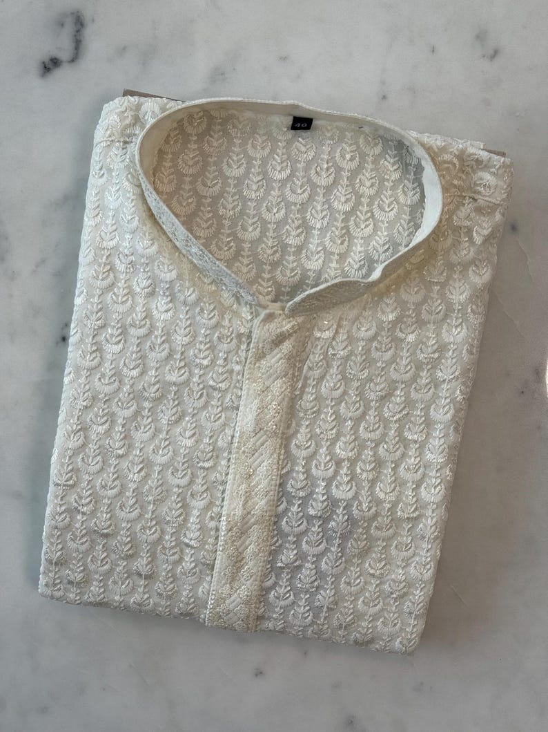 Pu&ograve; includere: Camicia kurta ricamata color crema con colletto alla coreana. La camicia presenta un motivo floreale all-over e una chiusura con bottoni. Il tessuto sembra leggero, adatto alle temperature calde. La camicia &egrave; piegata ordinatamente, mostrando i dettagli del ricamo.