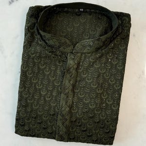 Pu&ograve; includere: Kurta ricamata verde scuro con colletto alla coreana. La camicia a maniche lunghe presenta un motivo floreale all-over e una chiusura con bottoni sul davanti. L'etichetta della taglia indica "40". L'indumento &egrave; piegato ordinatamente su una superficie bianca.