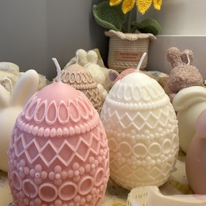 May include: Two decorative Easter egg candles, one pink and one white, with intricate lace-like designs. The candles are surrounded by other Easter decorations, including a crocheted sunflower and bunny figurines.