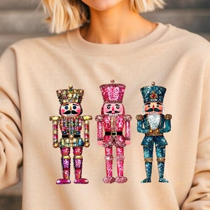 May include: A beige sweatshirt with a graphic of three nutcracker soldiers. The nutcrackers are decorated with sequins and have different colored outfits: gold, pink, and blue. The text on the sweatshirt reads "The design in the image is faux sequin. It is not real sequin."