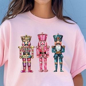 May include: A light pink t-shirt featuring three faux sequin nutcracker designs. The nutcrackers are in various colors, including pink, teal, and gold. The text at the bottom of the image reads: "THE DESIGN IN THE IMAGE IS FAUX SEQUIN. IT IS NOT REAL SEQUIN."