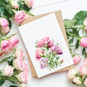 K&ouml;nnte beinhalten: Eine leere Gru&szlig;karte mit einem Aquarell-Blumenmuster aus rosa und lila Blumen, die auf einem braunen Umschlag liegt. Die Karte ist von rosa Rosen auf einer wei&szlig;en Holzoberfl&auml;che umgeben.
